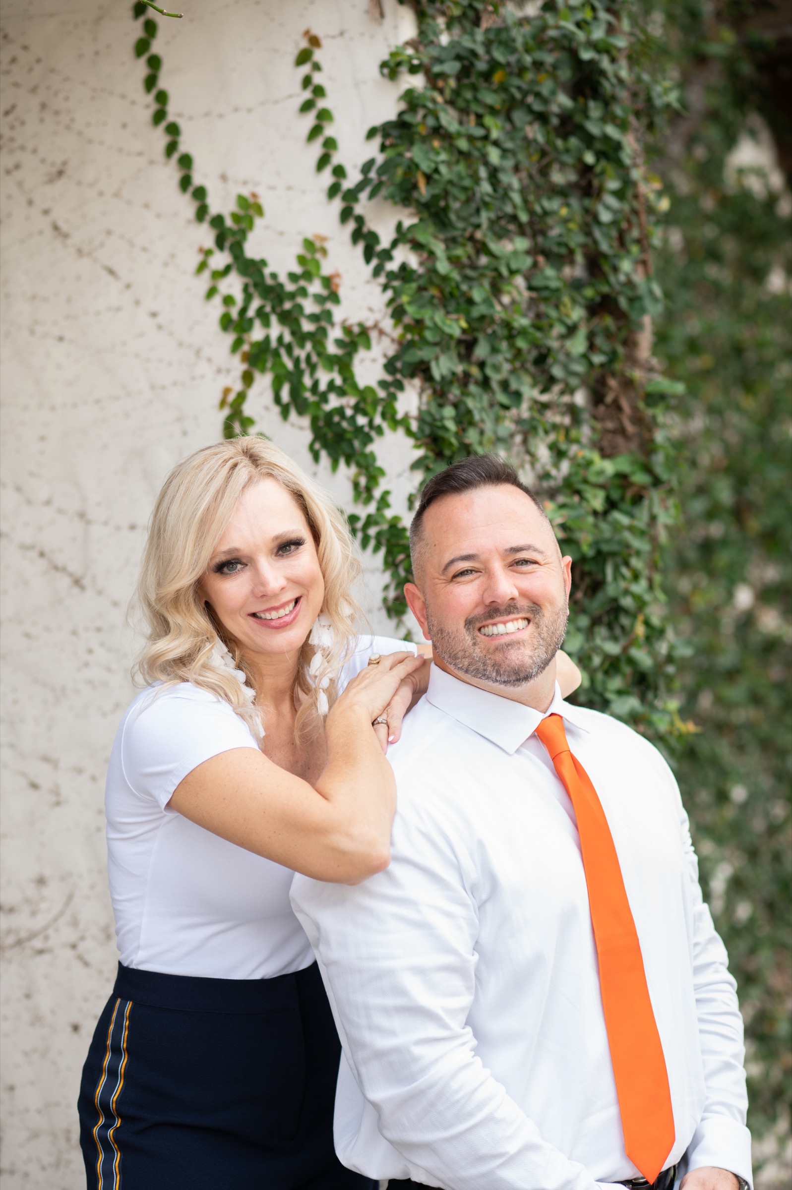 Jenn Freda resting a hand on Matt Freda's shoulder, both smiling, against an ivy-covered wall